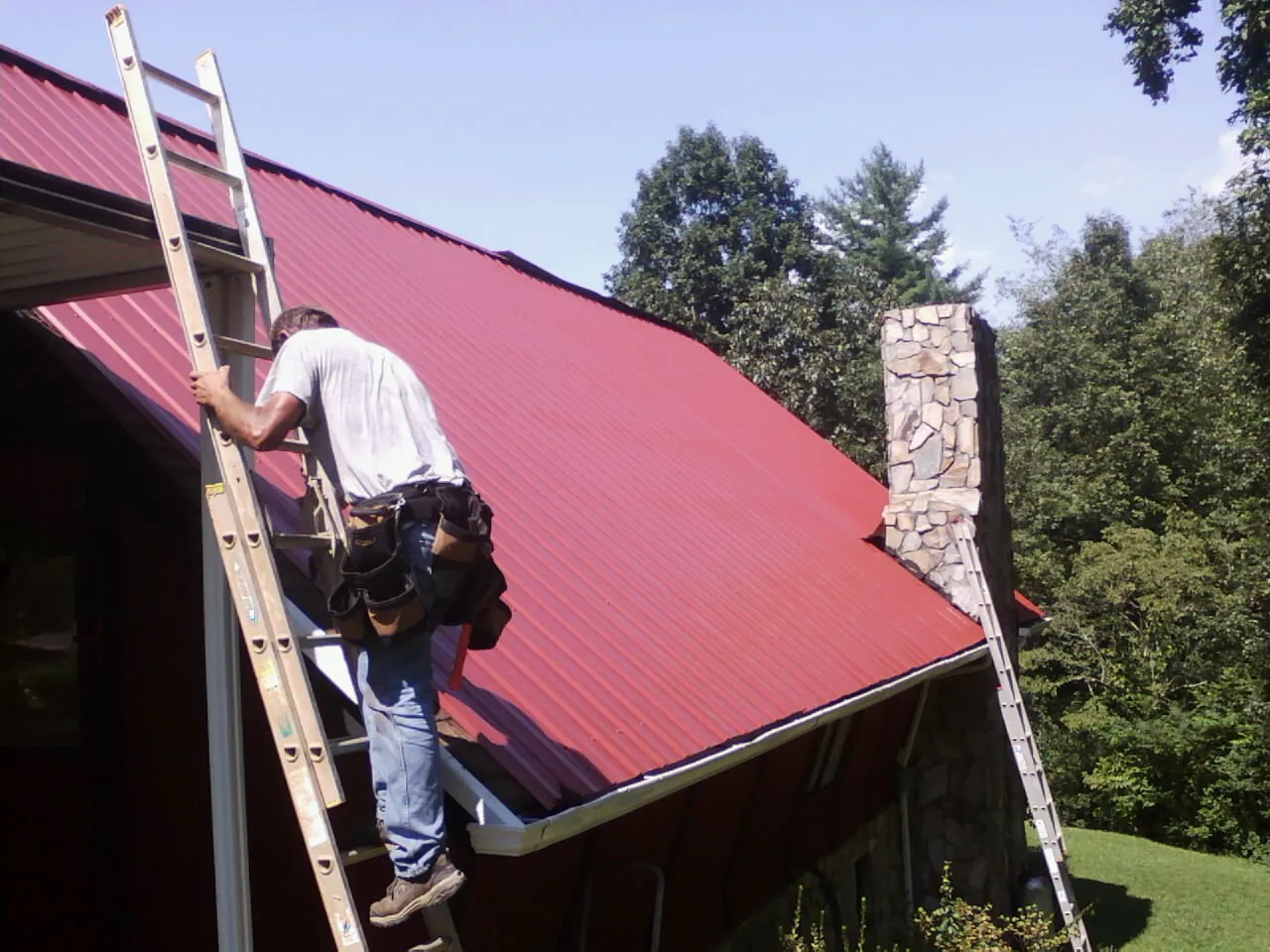 Roofer on a ladder beside a red metal roof and stone chimney — Soffit & Fascia Repair by Roofing Brothers in Buffalo Mills